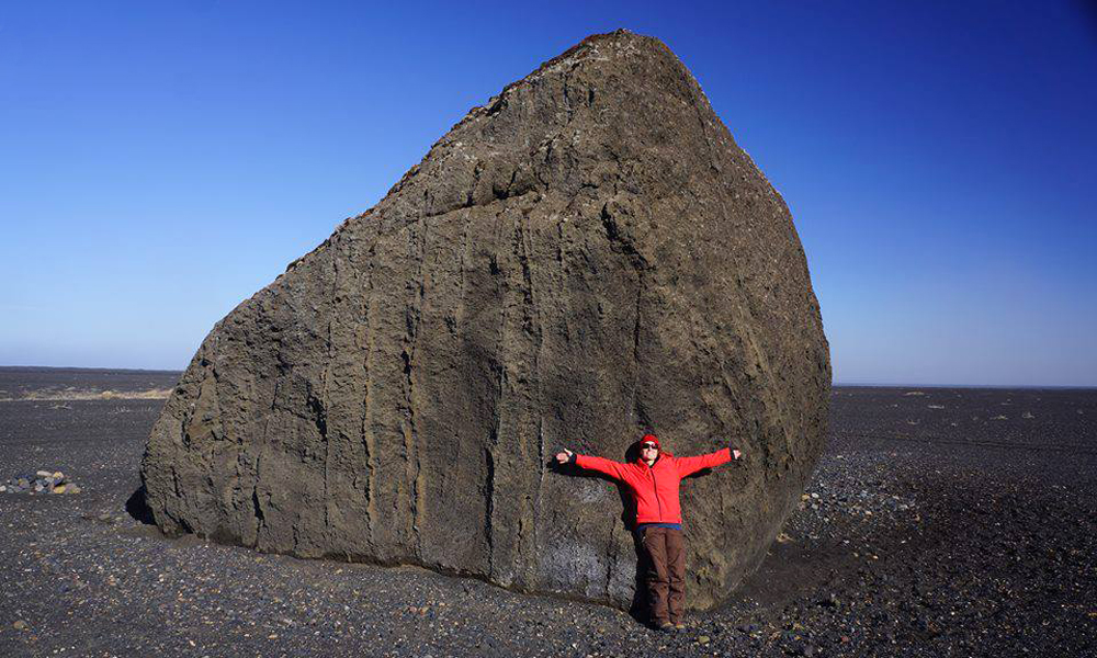 Erica Massey poses on a volcanic glacial outwash with a massive boulder