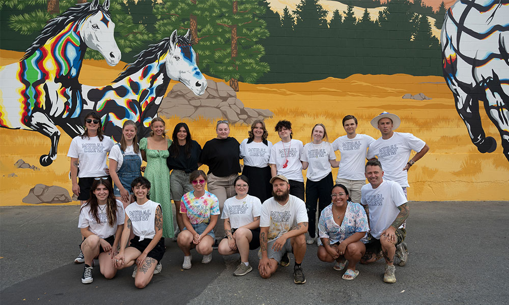 A group shot of 17 people standing in front of the 2023 mural. Some of the colourful horses in the mural can be seen behind the students in the background.