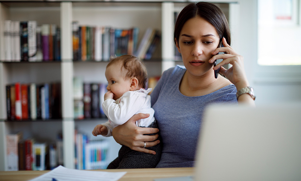 A woman looking stressed while trying to use the phone and carry a baby