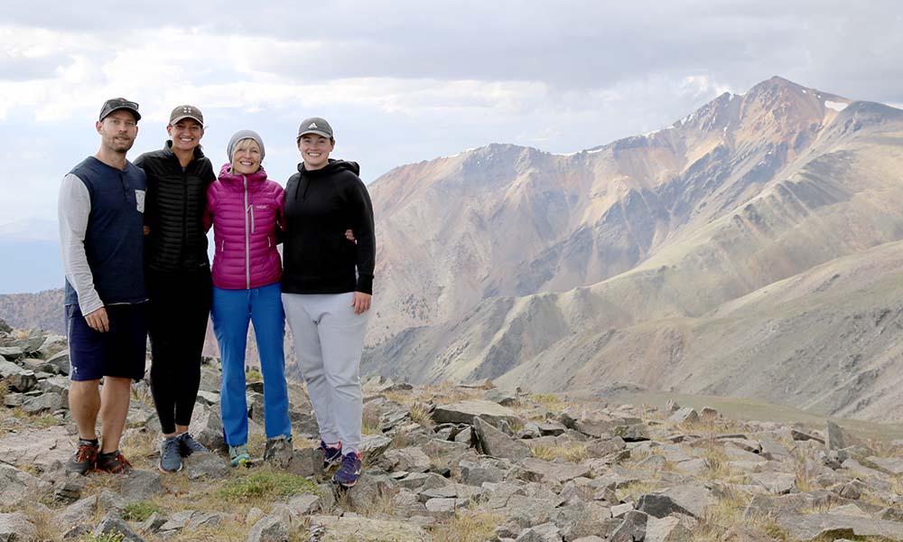 Four people standing at the top of a mountain, with an impressive mountain range seen in the background