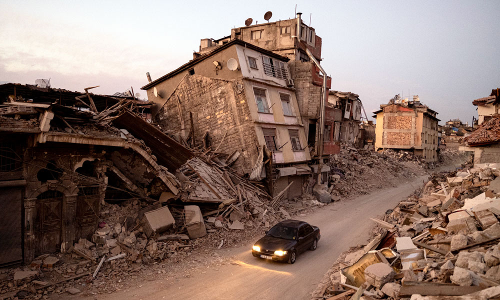 A car drives down a dusty road with buildings in the background devastated by an earthquake