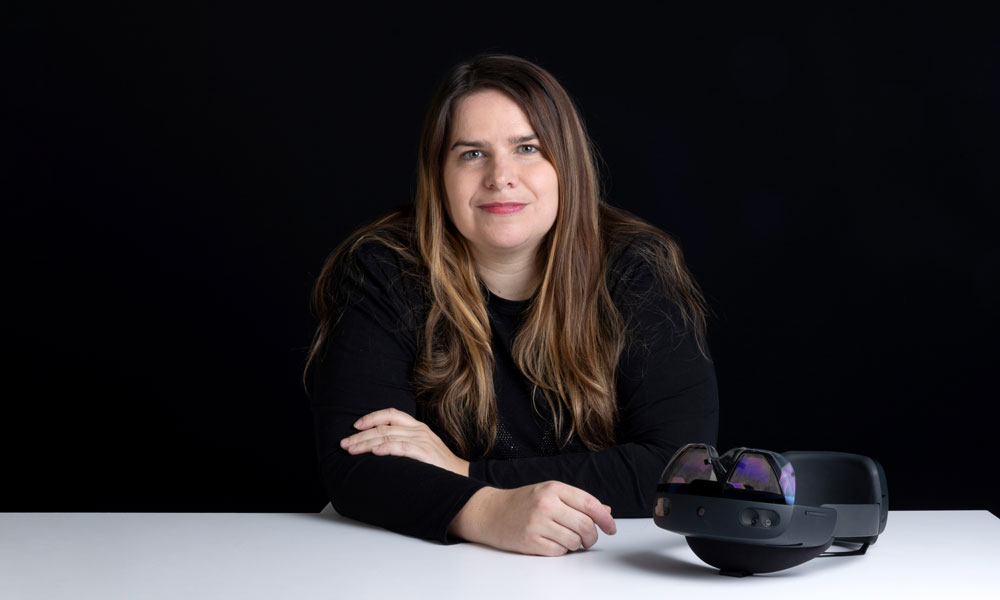 Carla Mather is dressed in black and leans against a white table, smiling with the microsoft hololens next to her on the table