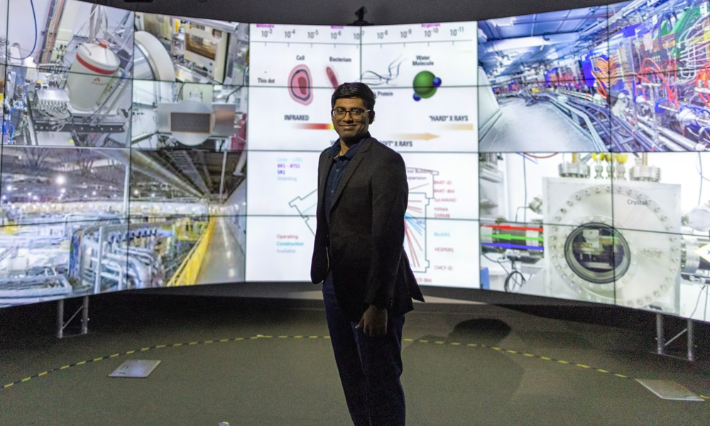 A photo of Bhavesh against the backdrop of the large TV screens, with scientific images on the screens behind him