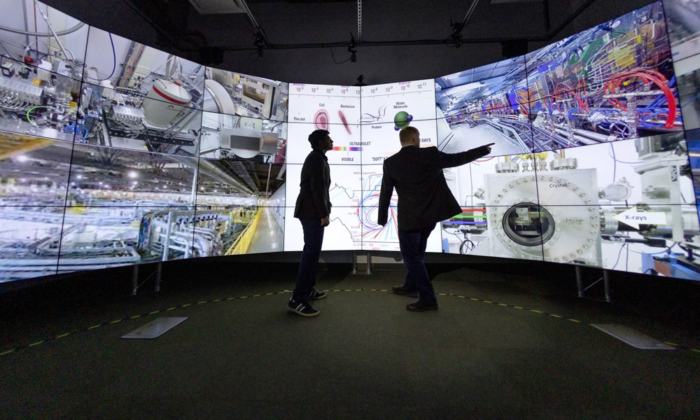 Bhavesh and Robert are shadowed by a giant rounded TV screen, which has a number of scientific-looking photos projected onto the screen