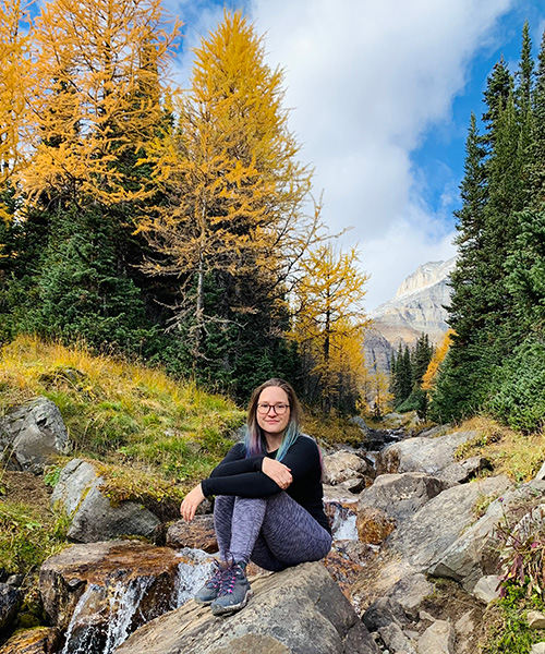 Melanie Dickie sitting on a large rock with a golden coloured tree behind her and blue sky