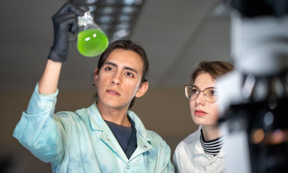 A man holds a beaker into the air while examining it. The beaker has a glowing green liquid inside it, and a girl stands next to him also looking at the beaker