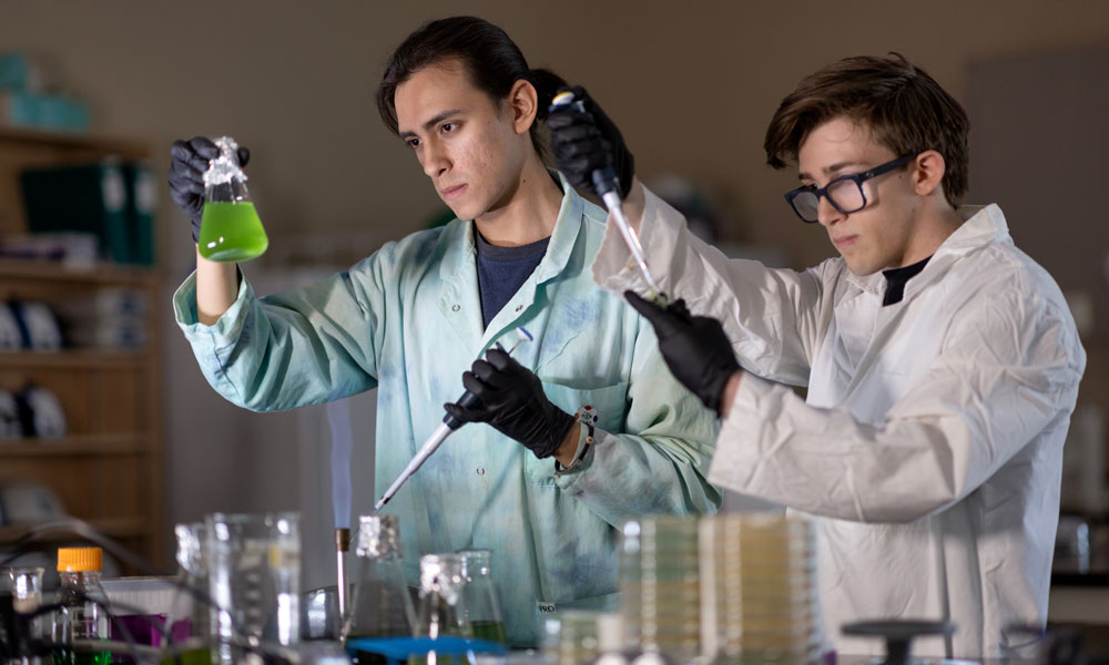 Two individuals in lab coats. one is looking intently at a beaker full of green liquid, while another is pipeting liquid into a test tube.