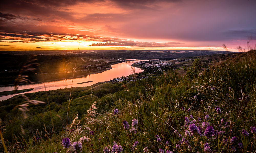 A photo of Peace River, with the sun setting in the background and a lush field in the foreground as a river snakes through the landscape.