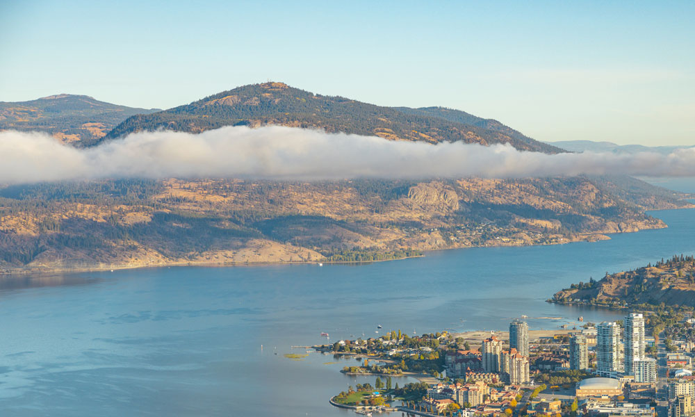 An aerial shot of Okanagan Lake, with a large mountain with fog in the background, and the city of Kelowna in the corner foreground