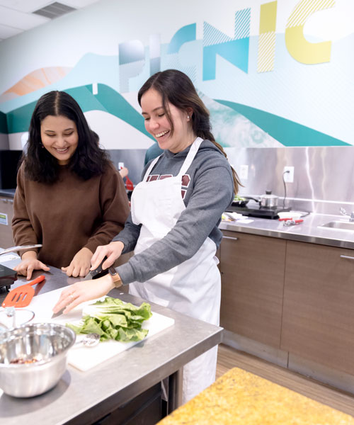 Two students laugh together while chopping bok choy, with the word "Picnic" seen above them on the wall