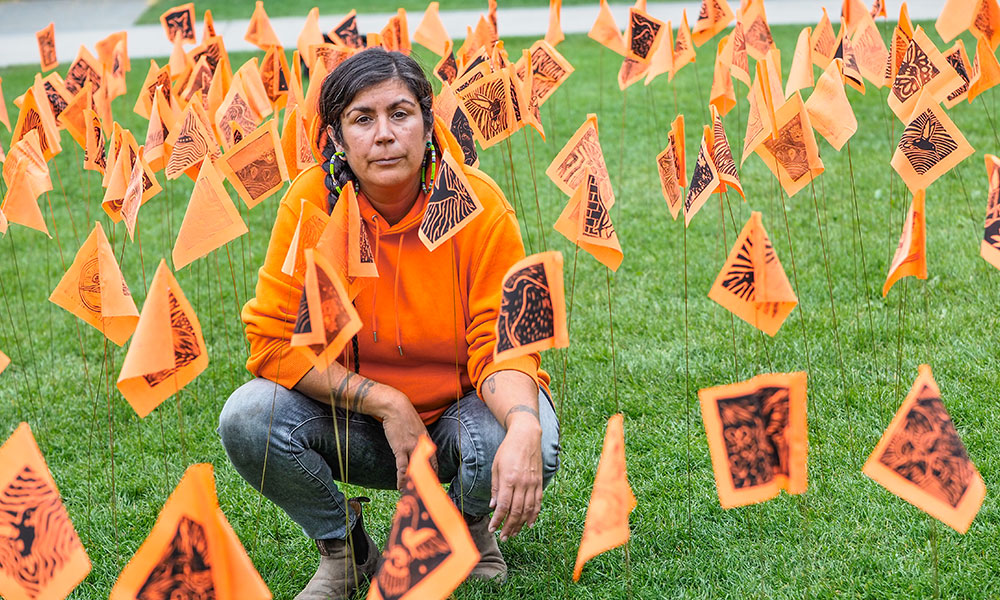 Tania Willard wears an orange sweater and kneels amongst dozens of orange flags inserted into the ground. Each flag bears a unique image of a hummingbird carved by workshop attendees.