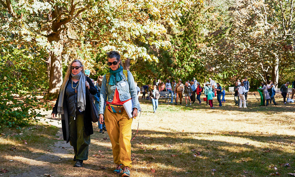 Dr. Astrida Neimanis walks in a autumn-coloured forest alongside another women. People are following the pair in the background.
