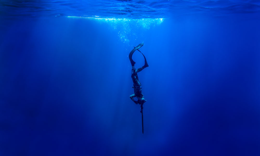 An underwater photo of a diver diving down into the depths of the ocean holding a spear-like object straight down. Bubbles and light rays of blue are seen at the top of the photo, signifying the surface