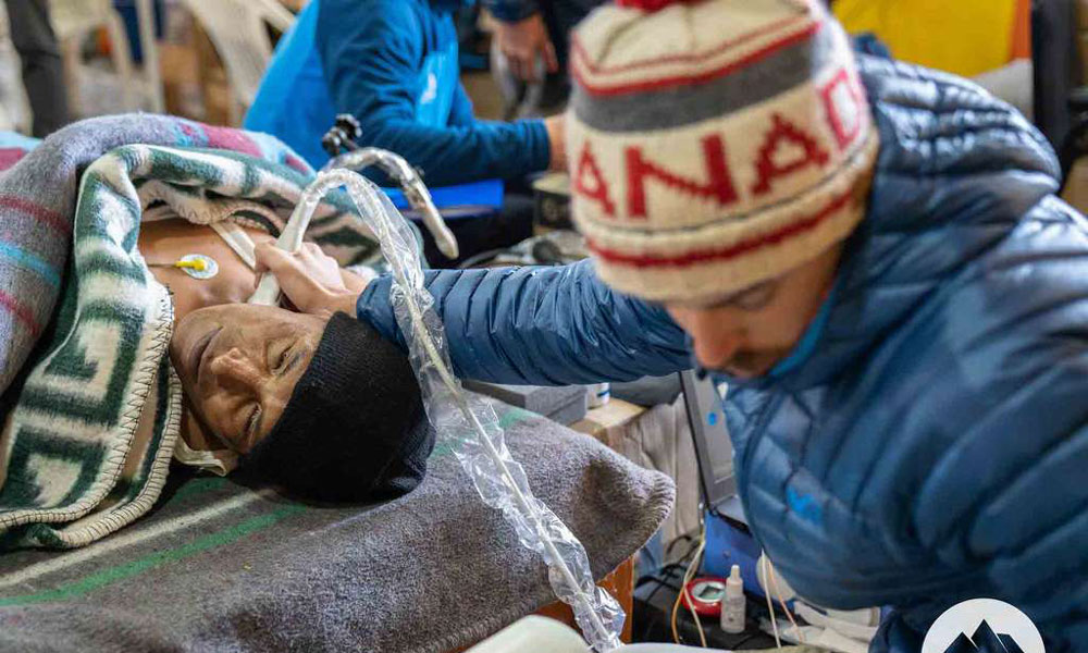 A man in a toque and warm jacket holds a ultrasound-type probe to a patient's neck. the patient is under a traditional peruvian blanket