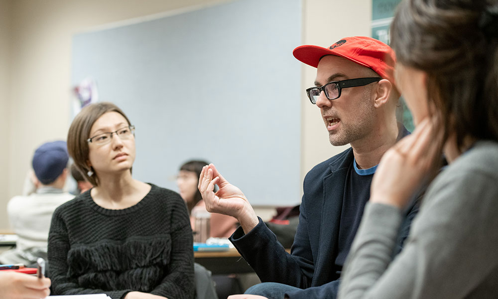 Michael V Smith is caught mid-sentence speaking as a student looks at him, intently listening