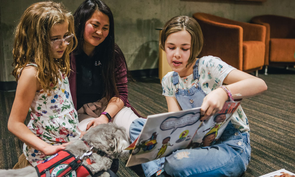 Camille Rouseeau looks on as a girl holds a book up to her sister and points to the page. A grey dog wearingh a BARK vest sits on the sister's lap while the girls read.