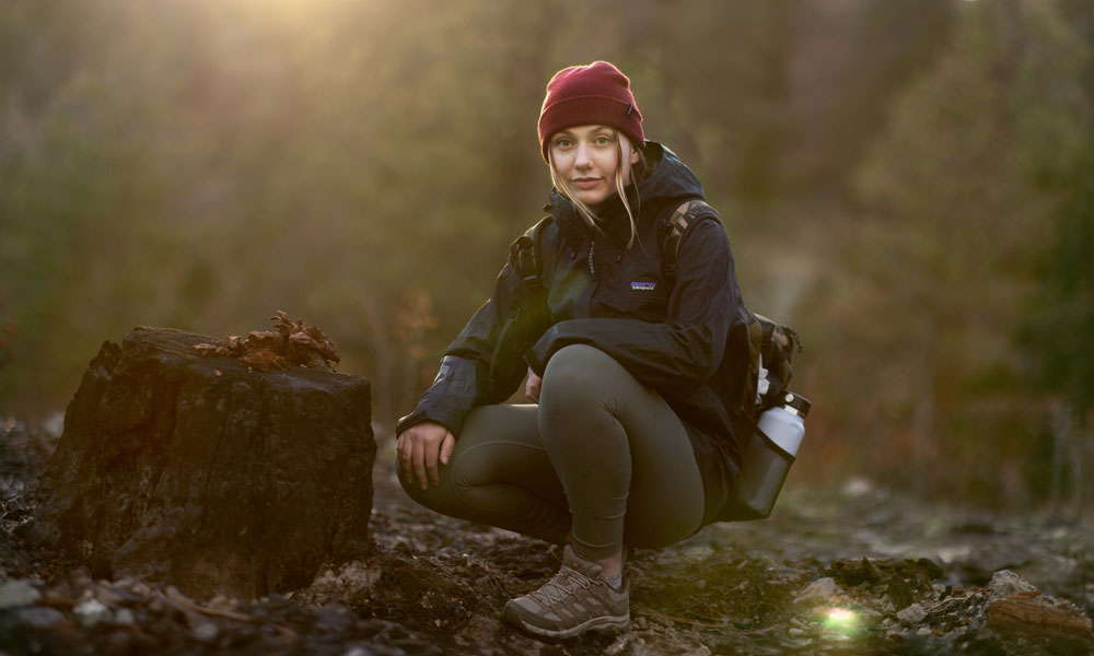 A young woman crouches next to a burned out stump in a forest, while the sun shines just over the mountain ridge in the background. The woman is wearing warm clothes and has a solemn look on her face.