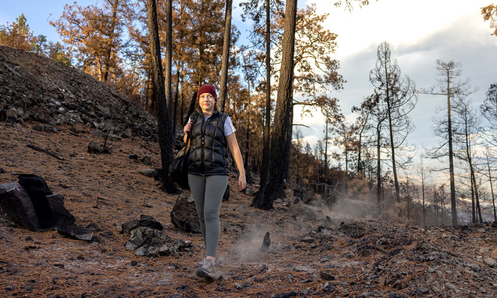A woman walks down a hill and kicks up ash behind her, a remnant from a recent forest fire in the wilderness she is in.