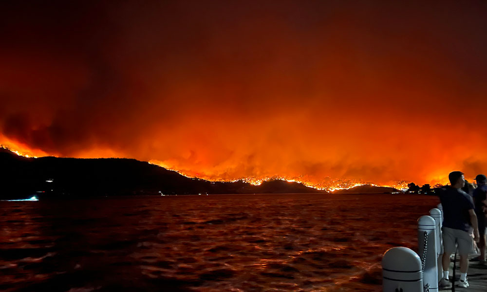 A man is seen on the edge of a photo standing watching a fire across Okanagan Lake that is burning an entire mountain side. The whole photo is a glow of orange from the flames.