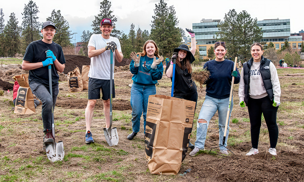 Six students pose with their gardening shovels as other students work away in the background