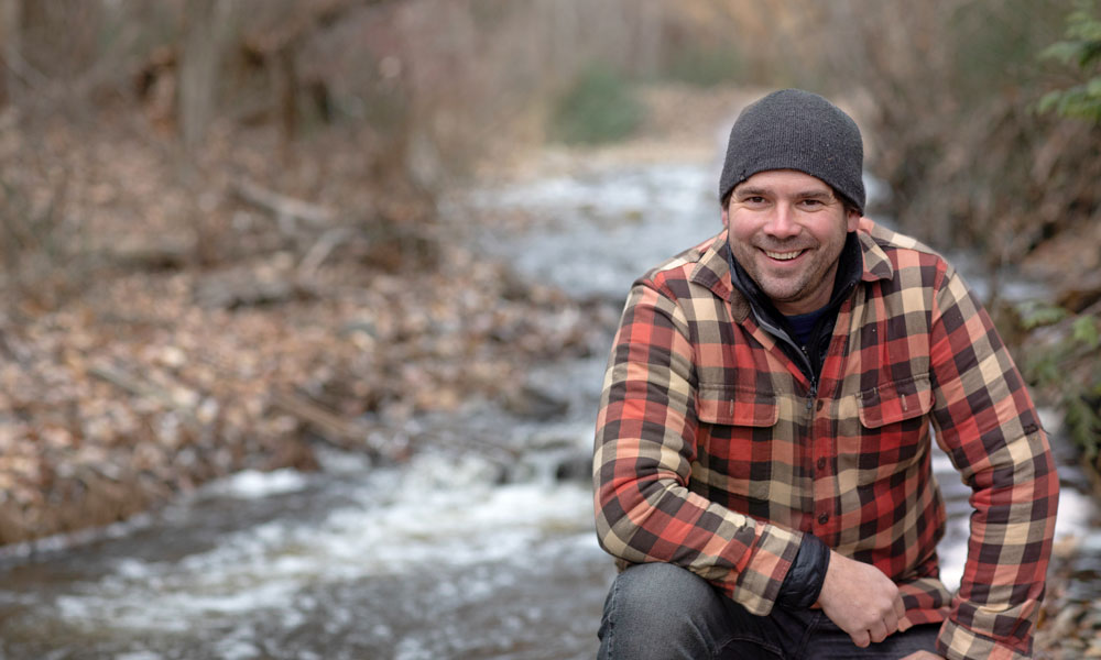Dr. Adam Ford kneels in front a creek, surrounded by trees and brush. He is wearing a toque and warm jacket