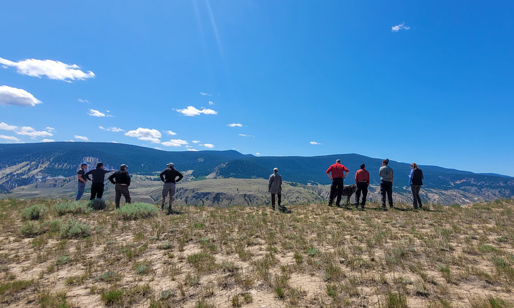A landscape photo of the backsides of nine people who are standing at the crest of a hill. The wide angle shot juxtaposes the group against the mountains in the background and blue sky above.