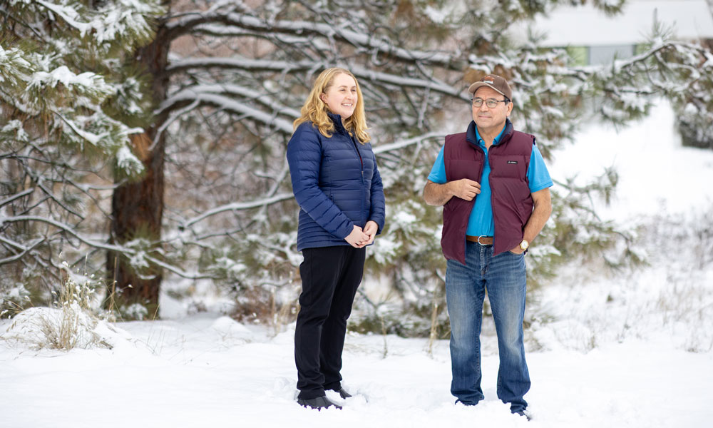 A photo of a woman and a man standing amongst snowy trees. They are both dressed warmly for the winter and are smiling as they look at nature.
