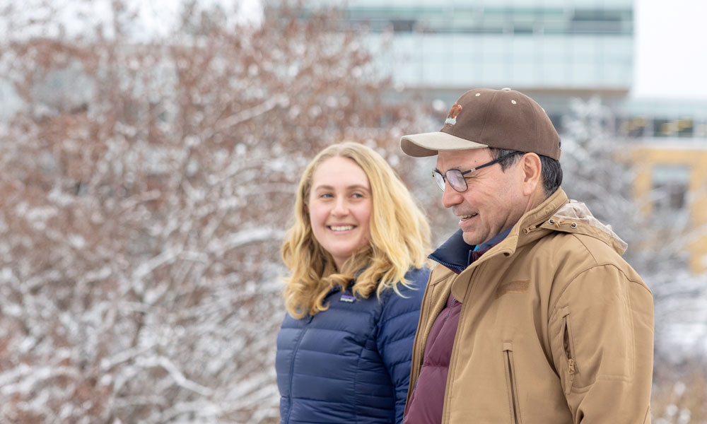 A blonde woman looks and smiles at a man while they both walk together on the UBC Okanagan campus. The Engineering, Management and Education building can be seen behind the two people.