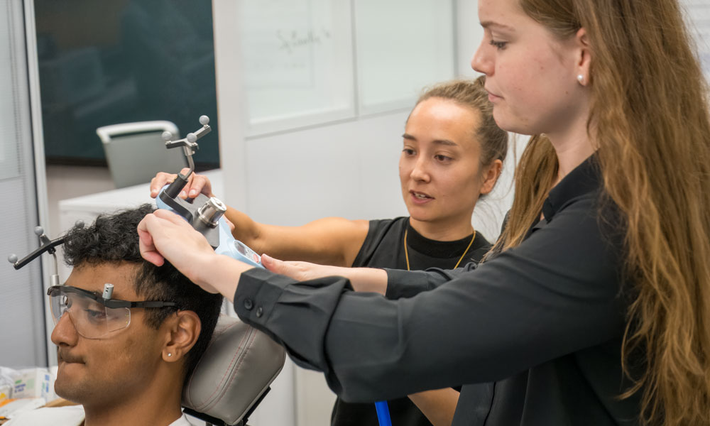 Dr. Sarah Kraeutner is featured in the background looking at a student seated with an apparatus on his head. Another student has her hands on the apparatus, adjusting it.