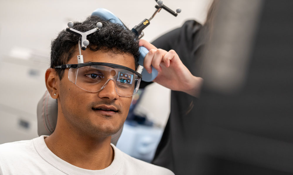 A close up of a student's face as he sits in a chair and an apparatus is attached to his head to measure brain function. 
