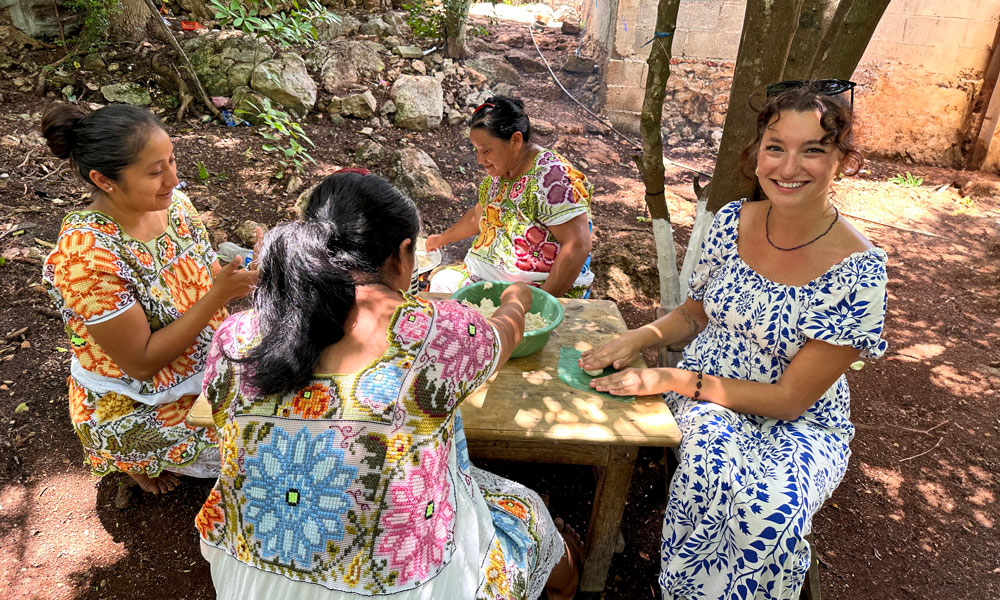 A student sits at a table outside, surrounded by three Mayan people. They are all making traditional corn tortillas while the Mayan people are wearing traditional Mayan embroidered dresses.