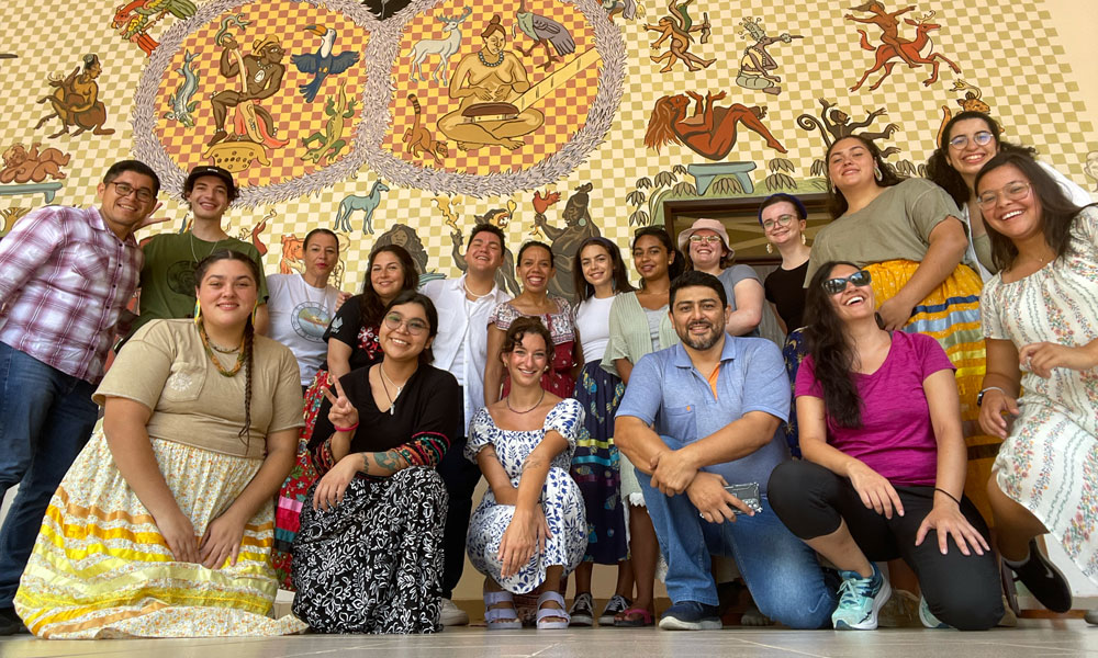 Approximately 20 people stand together for a group shot in front of a Mexican mural