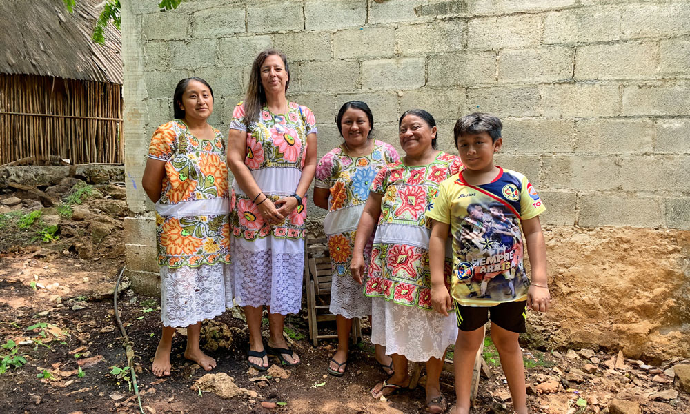 Five people stand in front of a brick wall, with four people wearing colourful embroidered dresses traditional of the Mayan people. Four of the people in the picture are from a Mayan community, while the fifth person is a visitor from Canada.