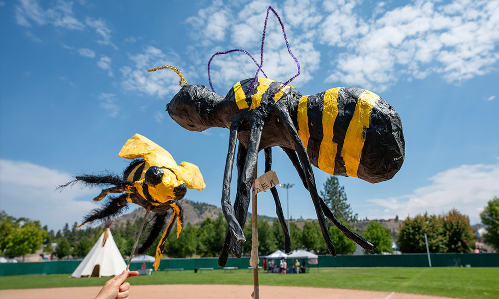 A close-up of two giant paper mache bees painted yellow and black, pictured against a blue puffy-cloud filled sky.