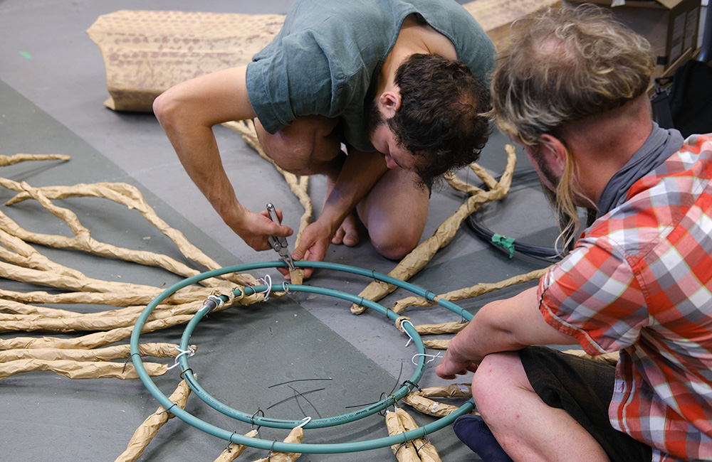 Two individuals are seen kneeling on the floor, trying to attach paper mache tentacles to two metal rings.