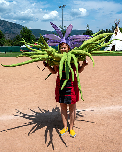 A woman stands on a sandy field wearing green tentacle-like strands around her neck, and a purple flower puppet attached to her head, representing a bitterroot.