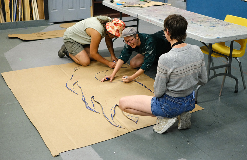Three individuals kneel on the ground and trace flames on a piece of cardboard. One person is seen laughing.