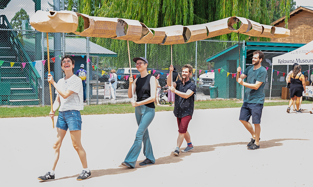 Four individuals hold sticks and at the top of the stick is a paper mache boa that is able to move and articulate as a community art piece.