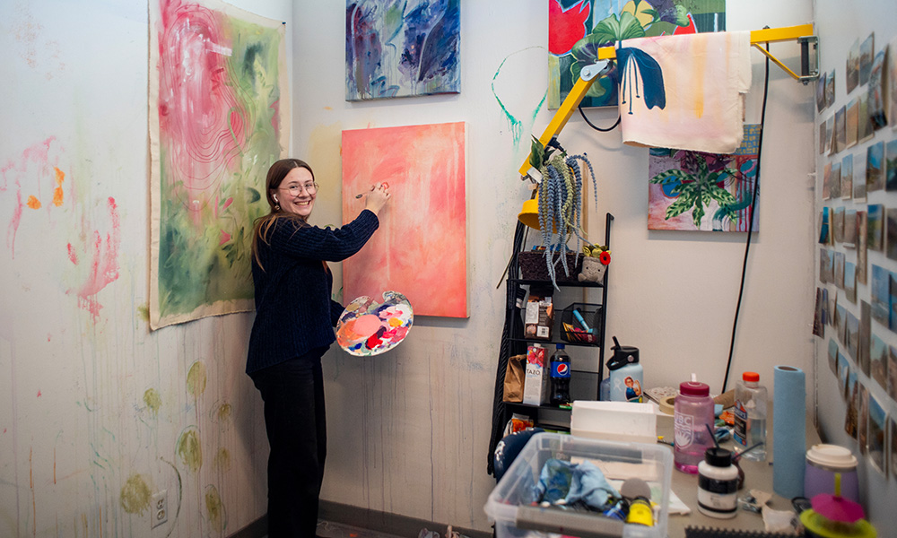 A photo of Bachelor of Fine Arts student Audrey Allen in an art studio surrounded by paintings.