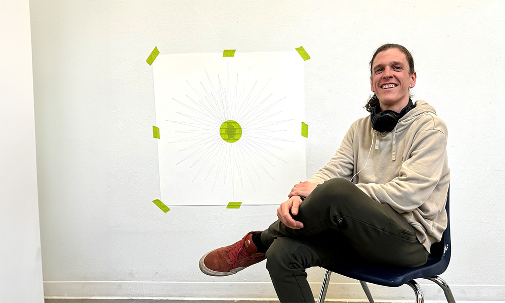 Asana Hughes sits on a chair positioned against a white wall. Taped on the wall with green painter's tape is a pencil outline of a mandala.