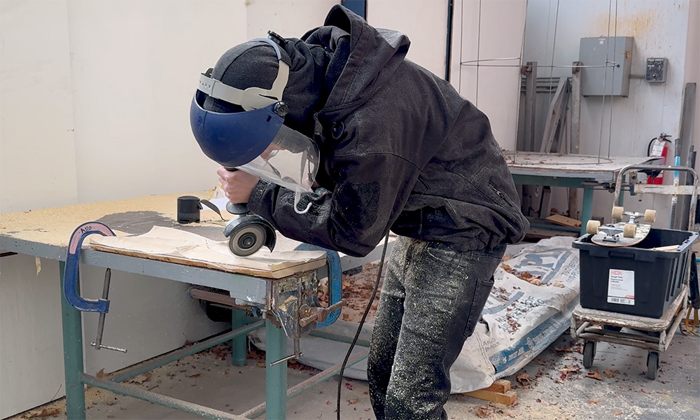 Asana Hughes in the metal shop, wearing a protective face shield while he uses a grinder on something placed on a table top.