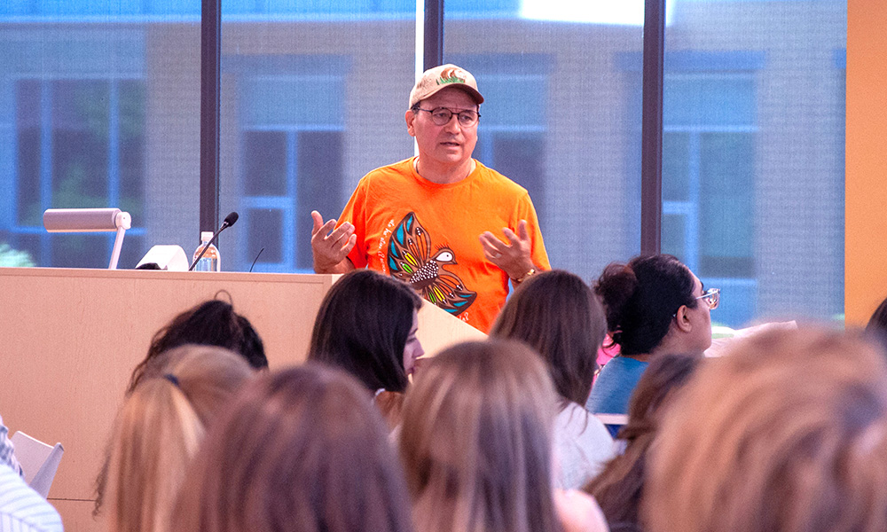 Dr. Bill Cohen stands in front of a classroom full of students, in the middle of explaining something. He wears an orange shirt with an Indigenous design on the front