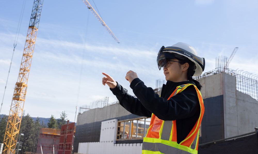 A woman stands on a construction site with her hands in the air, wearing a VR helmet. She points to something in the air, seeing it through her VR lens.