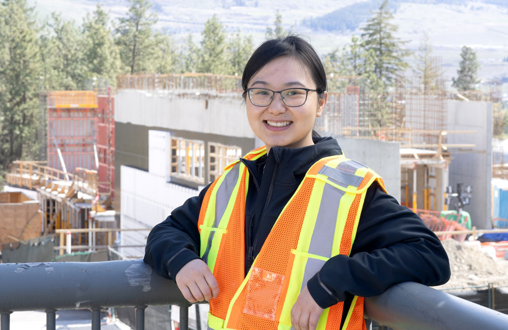 Dr. Qian Chen stands in front of a construction site, wearing a construction hi-viz vest, smiling and looking straight into the camera.