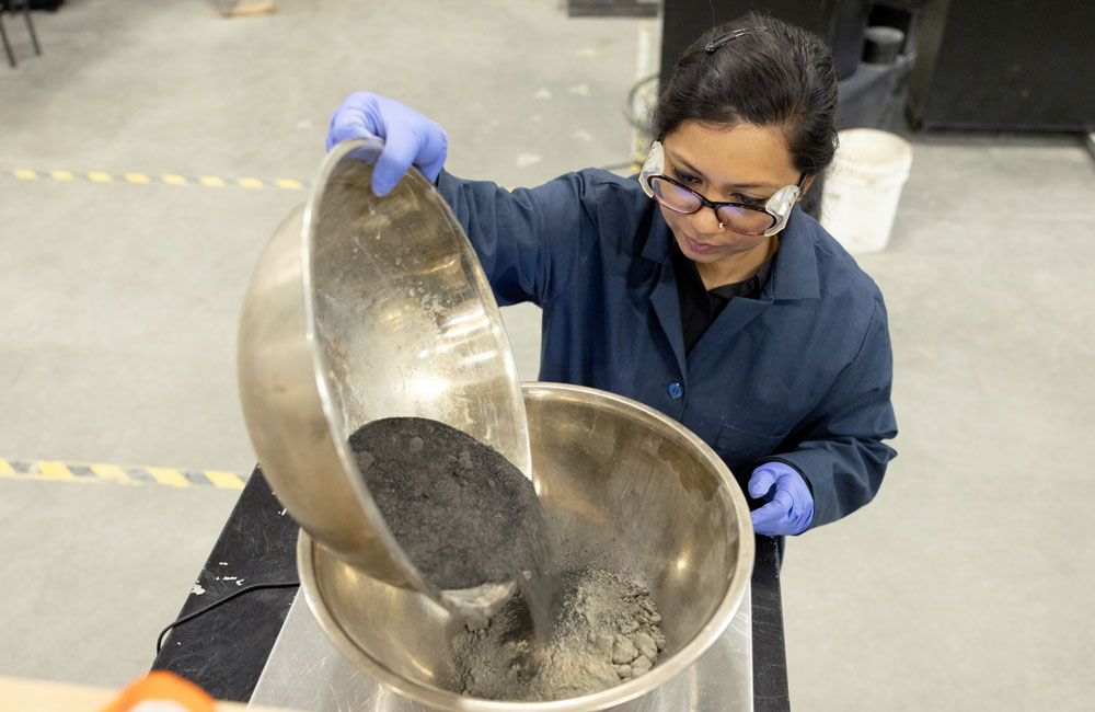 Taken from above, a woman in a blue lab coat wearing blue plastic gloves pours a grey dusty mixture from one large metal bowl to another, mixing the contents.