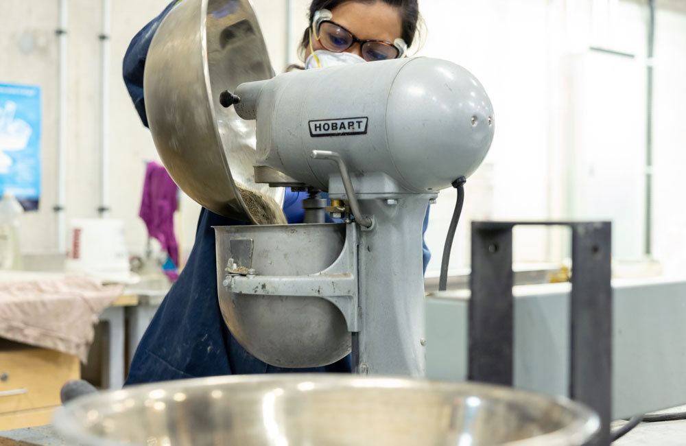 The woman pours the contents of the large mixing bowl to an industrial mixer.