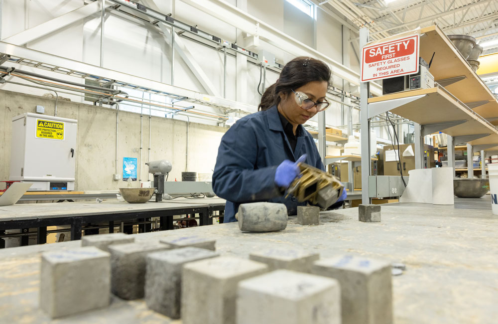 The woman in the blue lab coat is removing cement bricks from the mold in the background while the foreground shows a number of solid cement bricks.