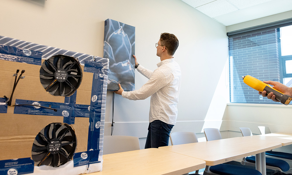 Jake Winkler hangs an ART device on the wall, while a second device is placed on the table nearby. The back of the device can be seen, including two large fans.