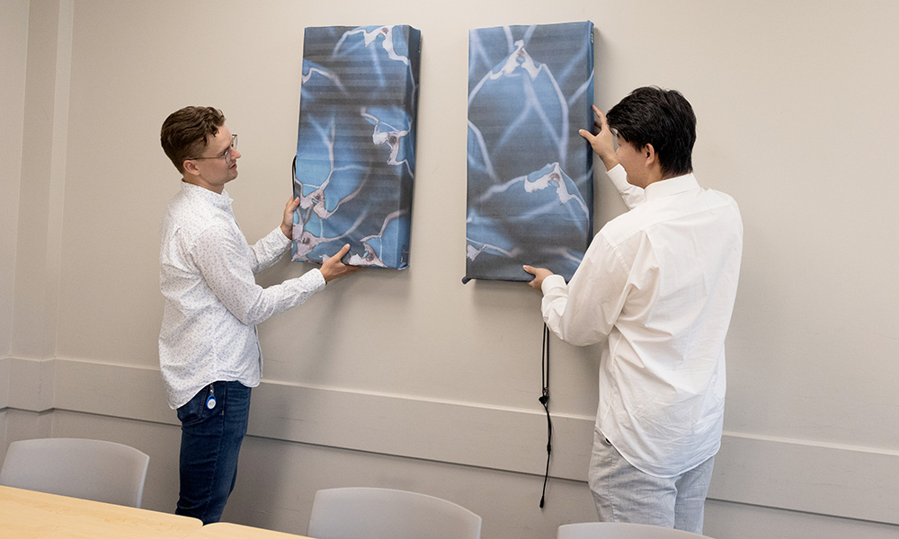 Jake Winkler and Xudong Wang each hang an ART device on the wall. The device looks just like an abstract painting with a small cord dangling from the bottom.