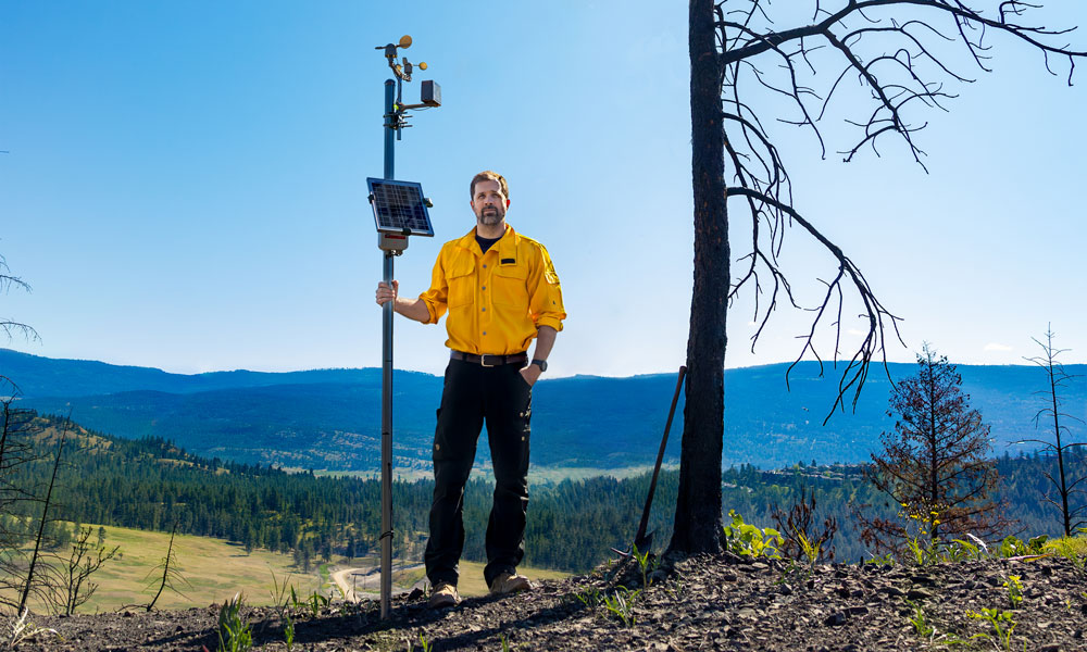 Dr. Mathieu Bourbonnais stands on a hilltop next to a burned, decaying tree. Wearing a yellow BC Wildfire Service shirt, Dr. Bourbonnais holds a wildfire sensor in his right hand. 